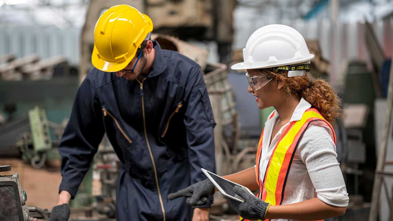 Female Machinery Engineer and male technical in safety protective wear while discuss with using tablet computer. They standing near CNC milling machine.