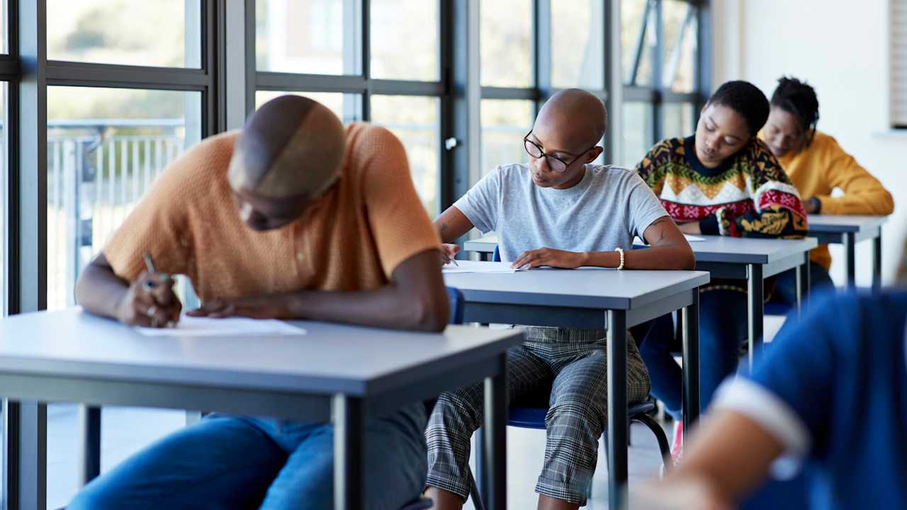 Young male and female university students writing on papers at desks by window in examination hall