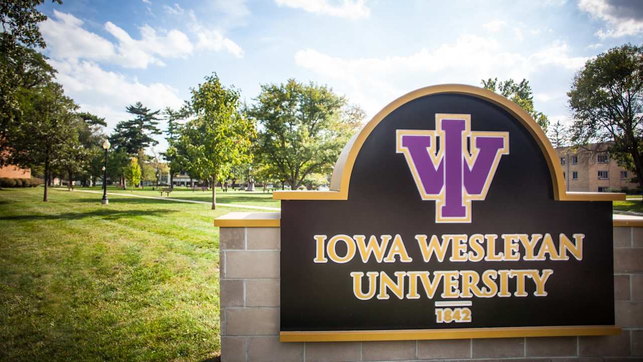 A picture of a Iowa Wesleyan University sign. The sign is photographed against a background of trees and grass on a clear sunny day