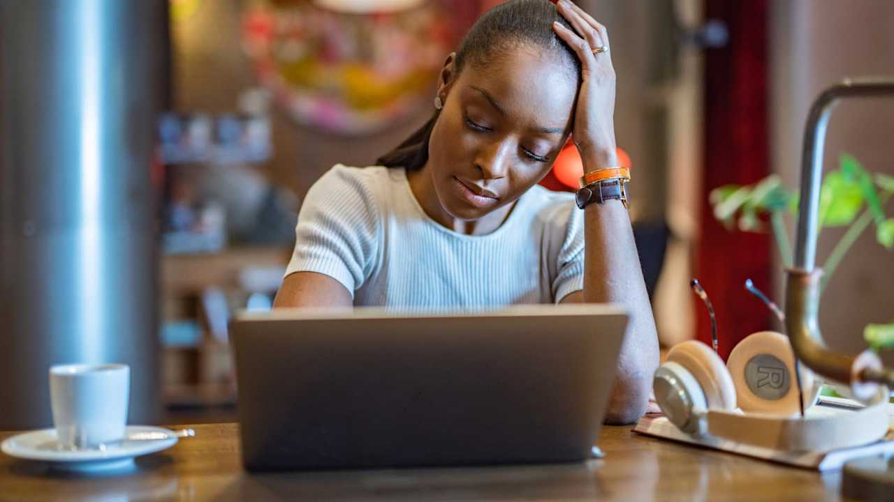Tired college student taking a break from typing on her laptop computer. She is sitting in a cafe, and is resting her left hand on her head with her eyes closed.