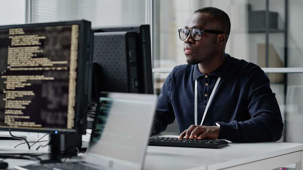 Black male cybersecurity engineer sitting at his desk in a corporate office. He is typing code for an internal security program on his desktop computer.