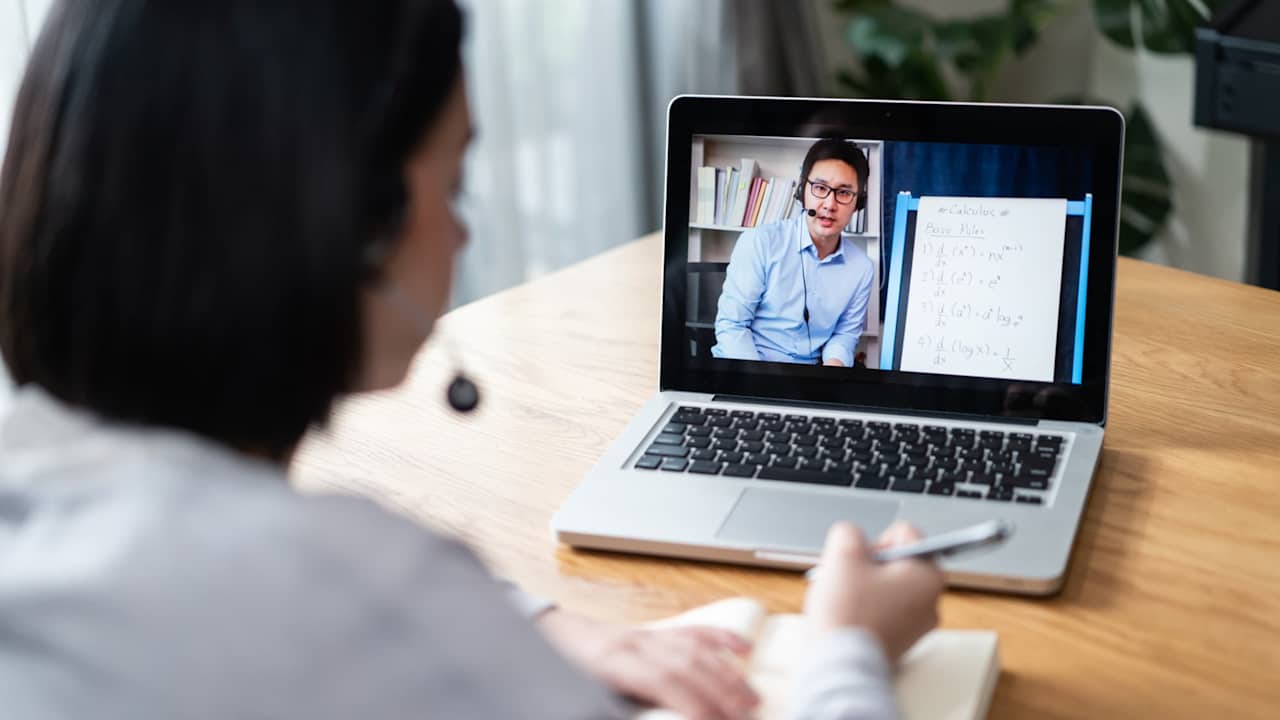 Student sitting in her home living room with her laptop open is taking an online calculus class for college. She is watching her instructor on her laptop screen demonstrating calculus equations on the whiteboard behind him.
