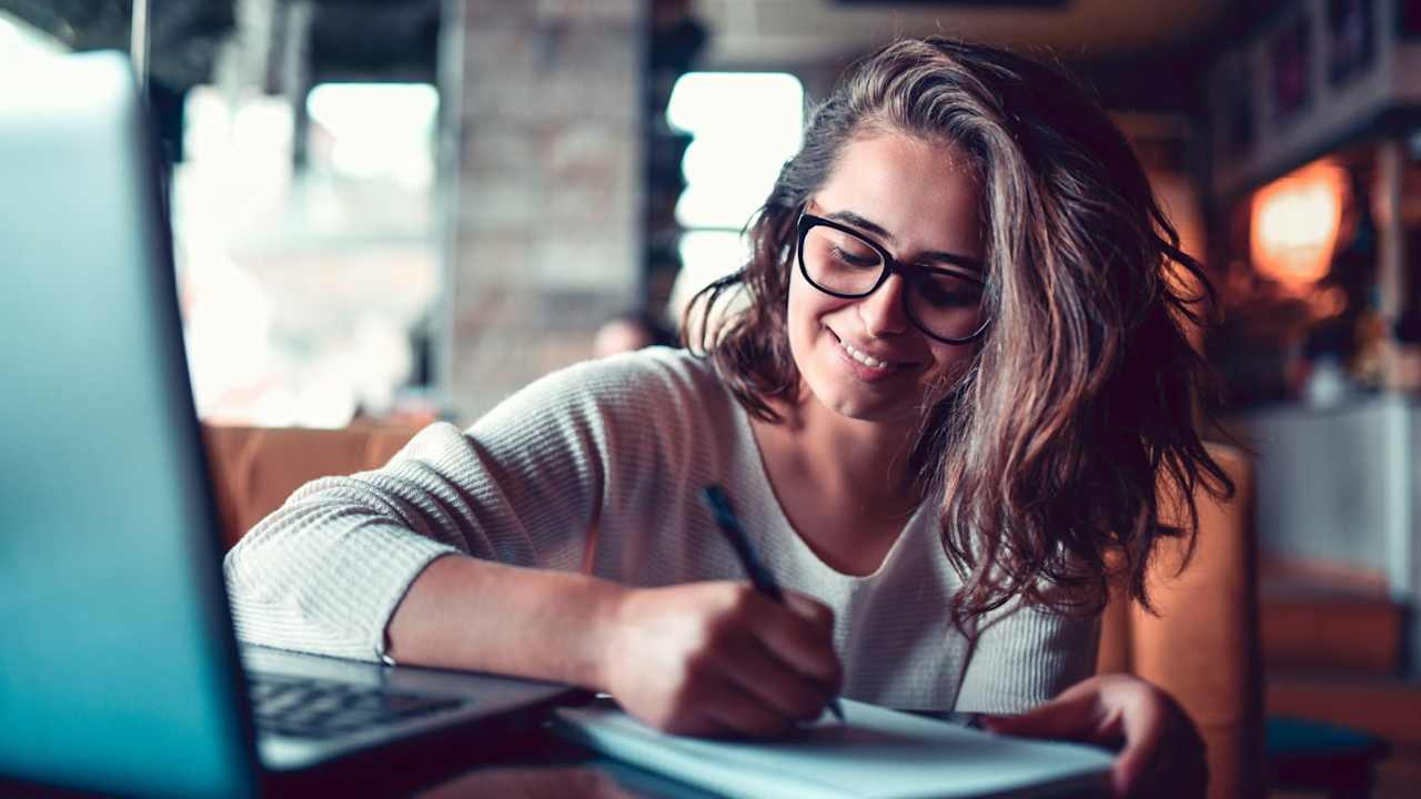 Female Taking Notes For Work Project While Researching Topic On Laptop
