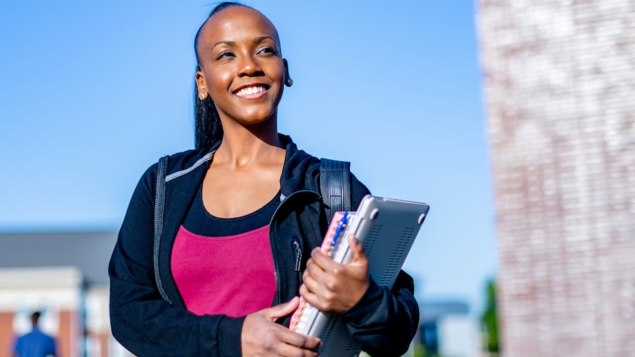 Black female college student smiles while walking to class on campus. She is wearing a backpack and carrying a notebook and laptop in her hands.