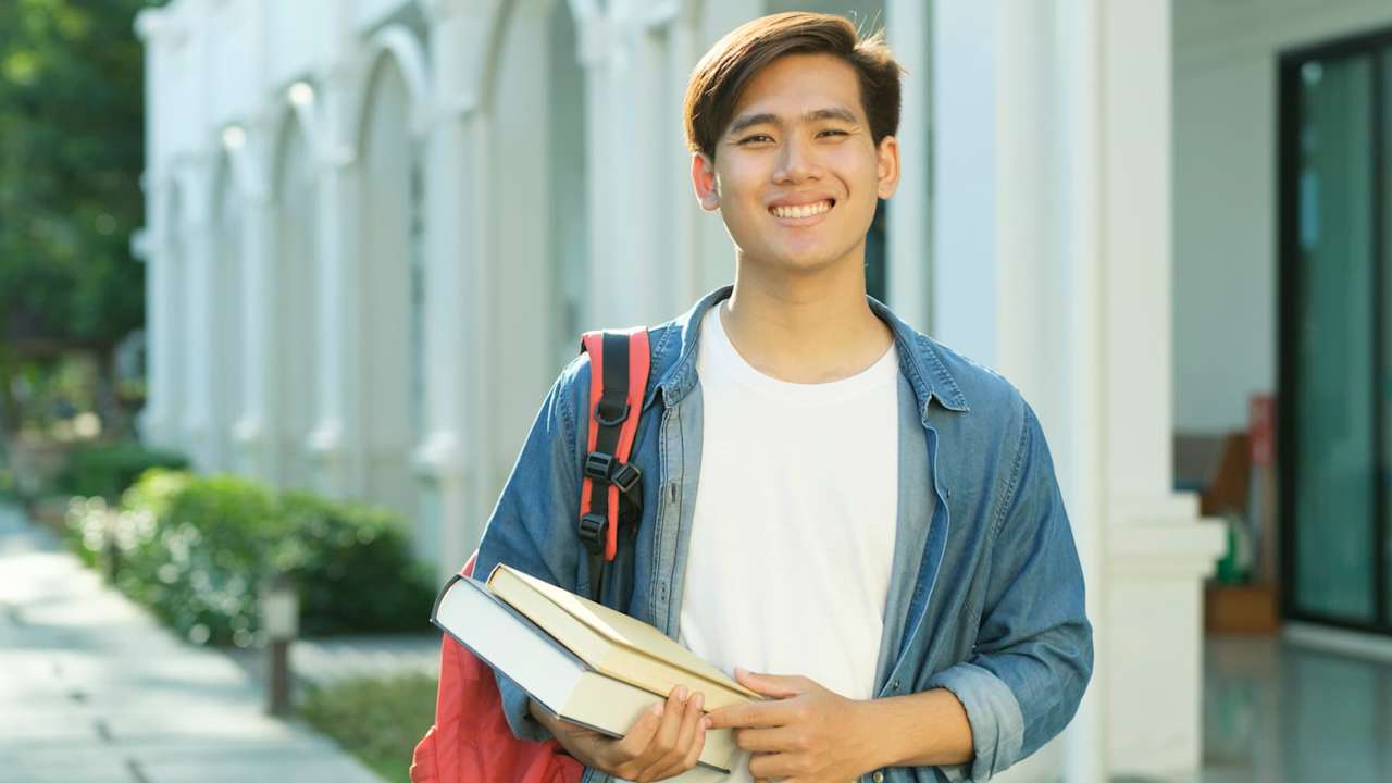 Male Asian American college student wearing casual clothes and a backpack smiling towards the camera. He is holding textbooks while standing outdoors on campus.