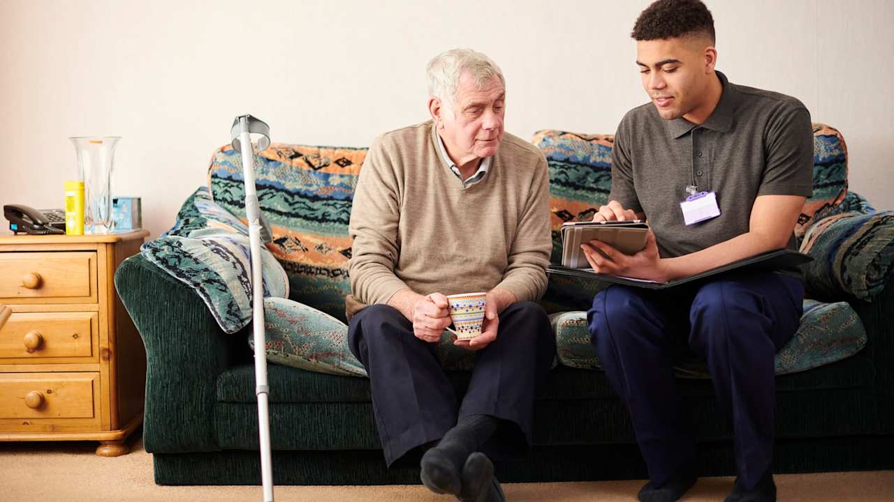 Black male social worker paying a house visit to a senior male client. They are sitting on the client's couch in his living room and chatting together.