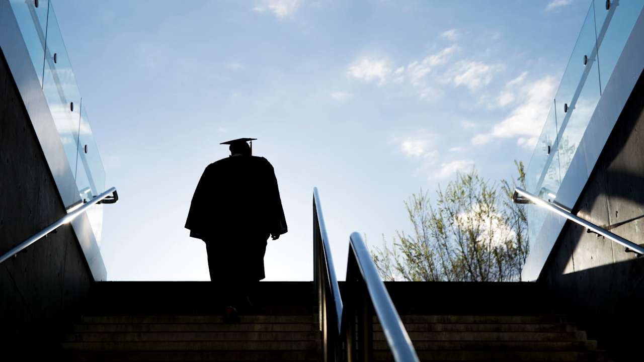 Silhouette of a college graduate in a cap and gown climbing up a set of steps on a university campus.
