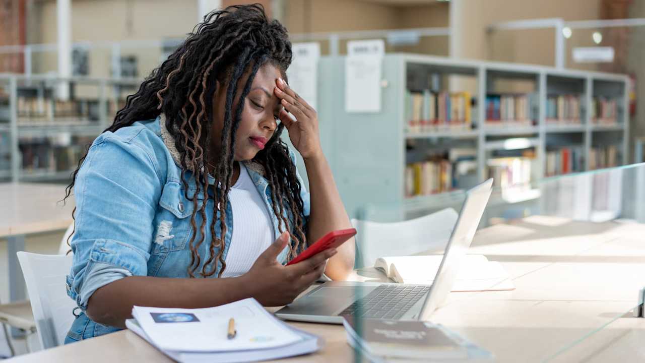 Black female student applying to college on her laptop. She is resting her head on her left hand, with a frustrated look on her face.