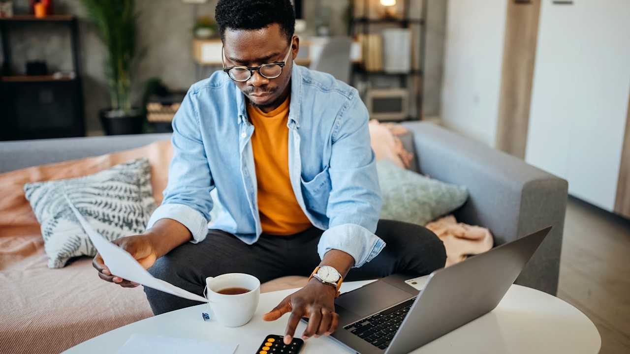 Black male college student sitting on a couch in his living room. He has his laptop open on the coffee table, and is holding a paper bill in his right hand. He is typing numbers on a calculator to check the bill amount on his student loan bill.