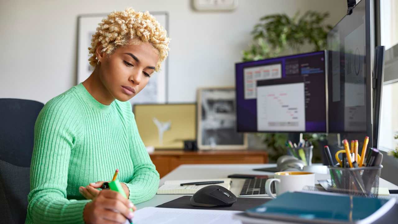 Black female business professional working from home at her desk. She is writing notes with a felt tip pen on paper while looking at charts and spreadsheets on her desktop computer.