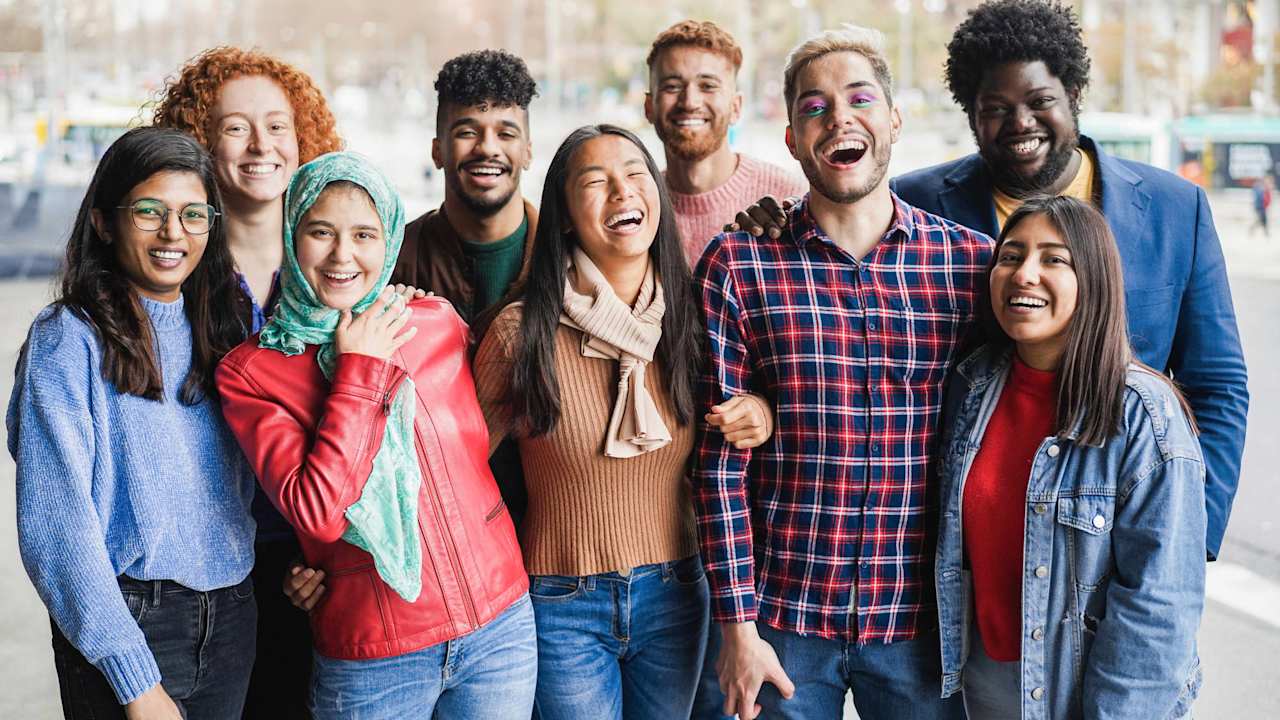 A diverse group of male, female, and non-binary college students standing together outdoors on campus, laughing and smiling with each other.