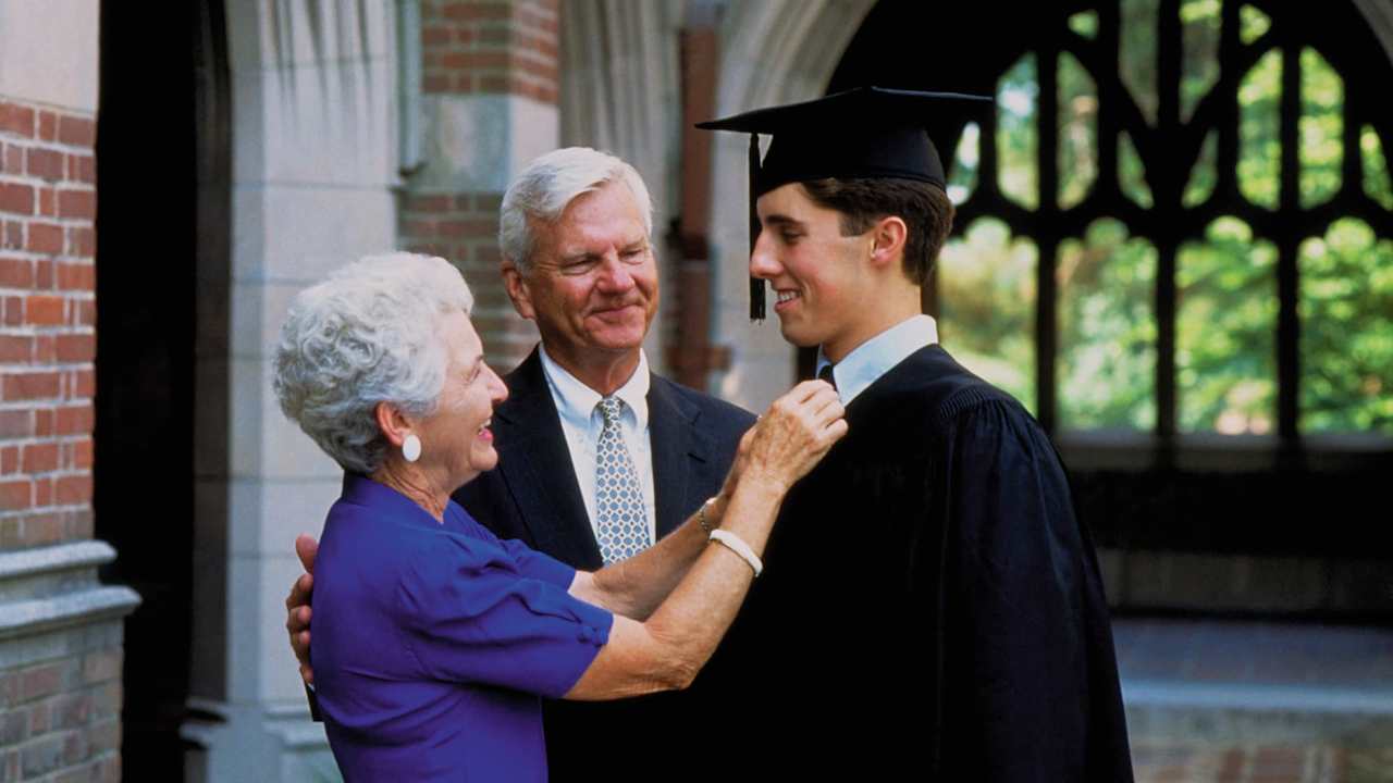A Caucasian male college student dressed in a black cap and gown celebrates with his grandparents on his graduation day. His grandmother is adjusting his tie, while his grandfather gives him a congratulatory pat on the back.