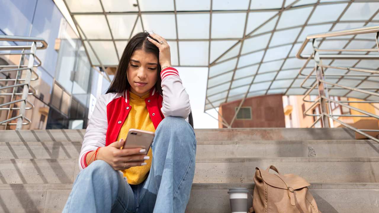 Latina college student sitting on steps outside of a building on campus. She is reading on her cell phone, while resting her arm against her head, with a stressed look on her face.