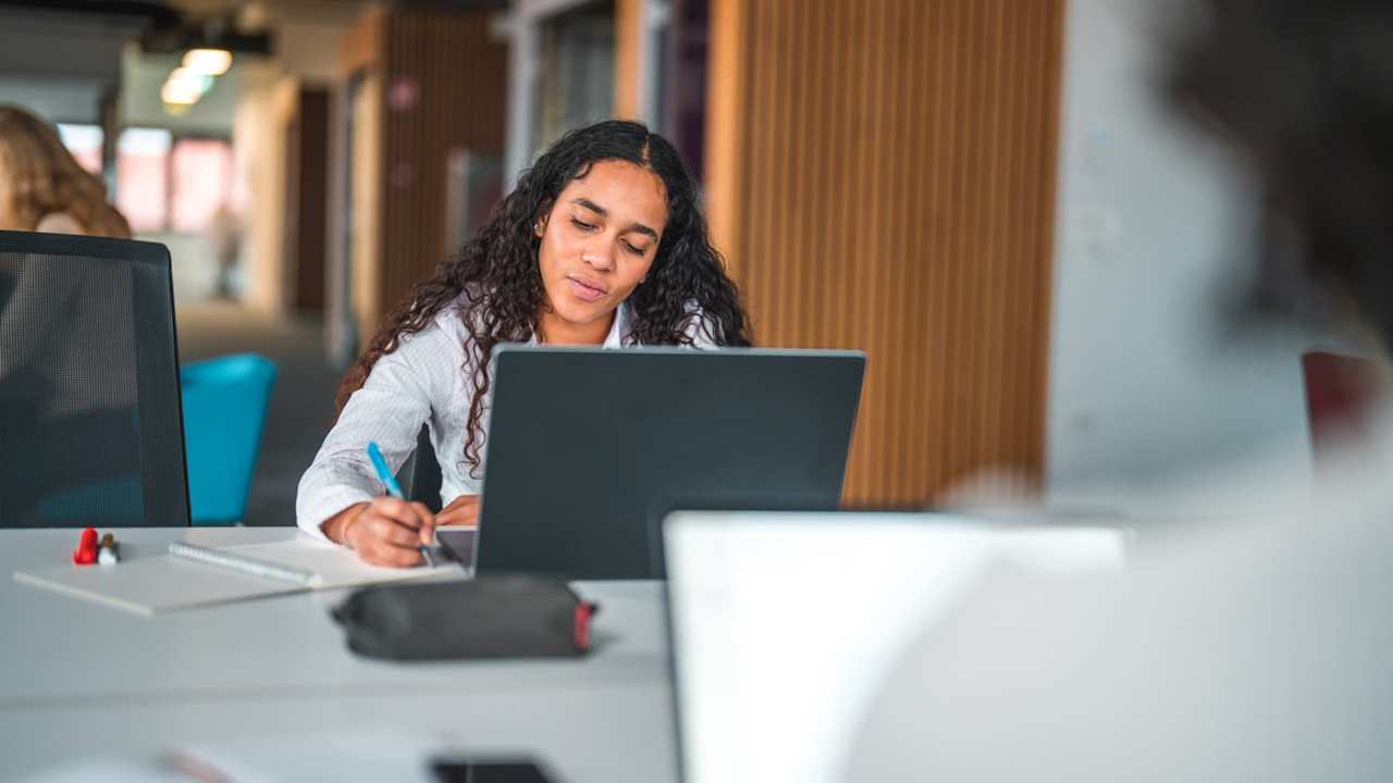 Female South American employee working on a laptop, with a female coworker sitting across the desk from her.