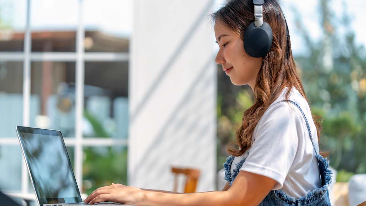 Female Asian American college student sitting at an outdoor table on campus on a sunny warm day. She is wearing headphones and typing on her laptop.