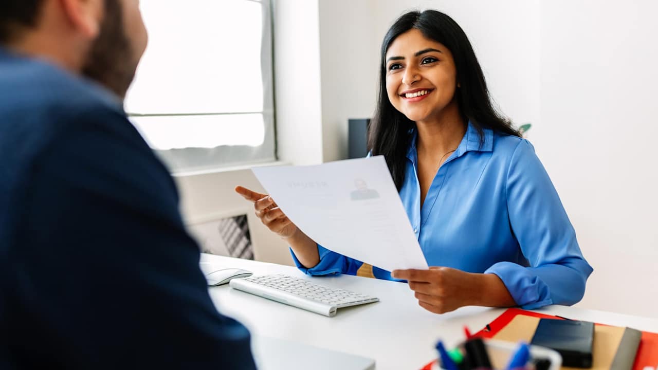 Indian female HR recruitment manager holding a paper resume in her hands while having an interview in a modern office. She is looking and listening to a Caucasian male job candidate, pictured offscreen.