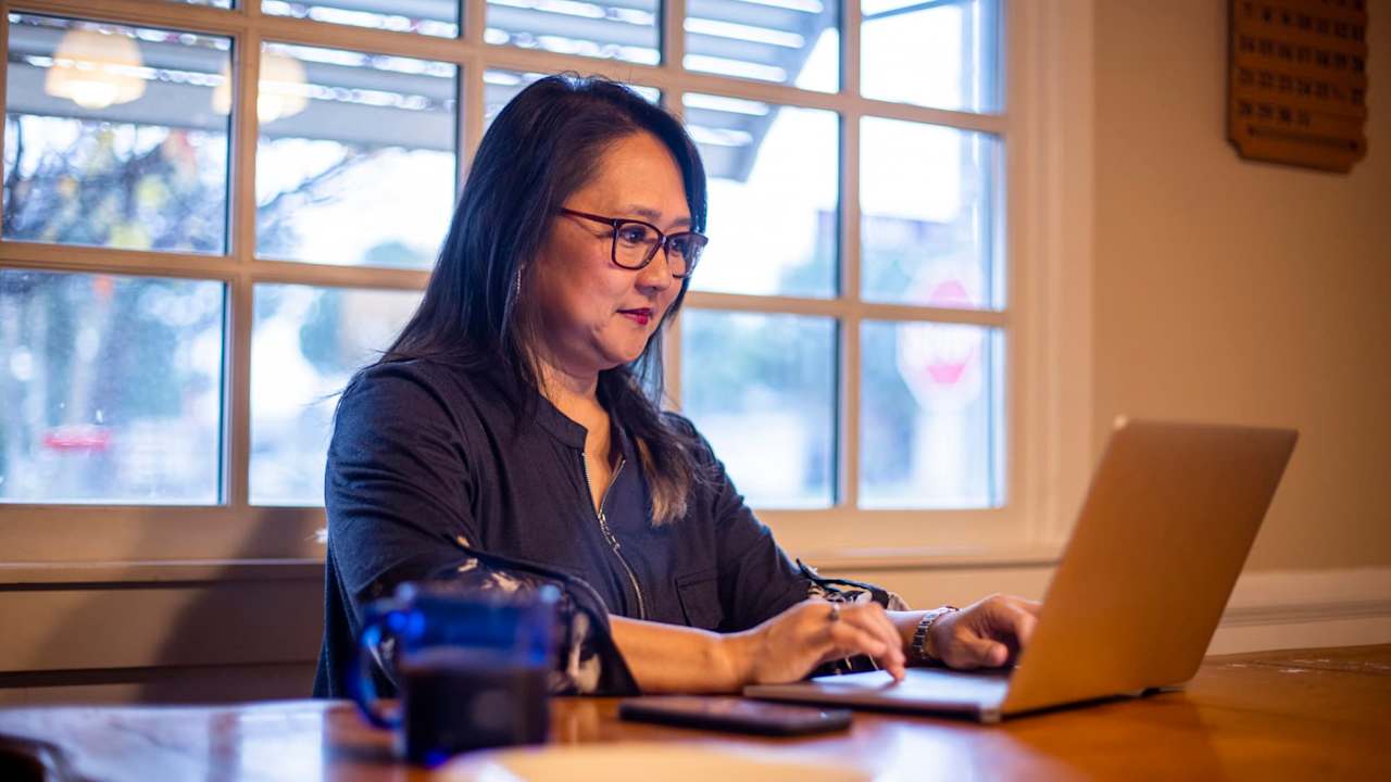Asian American female business student in her mid-40s typing an assignment for her online class at home. She is sitting at her dining room table, and is wearing glasses and business casual clothes.