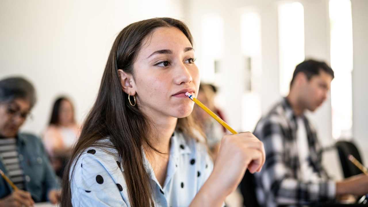 Latina college student sitting in a classroom with other students taking an exam. She has her head up and is putting the eraser tip of her pencil to her mouth while looking deep in thought.