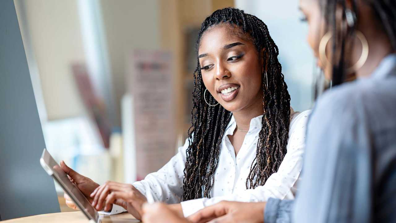 A Black female business development lead in a discussion with her female teammate via a tablet computer in a tech business office.