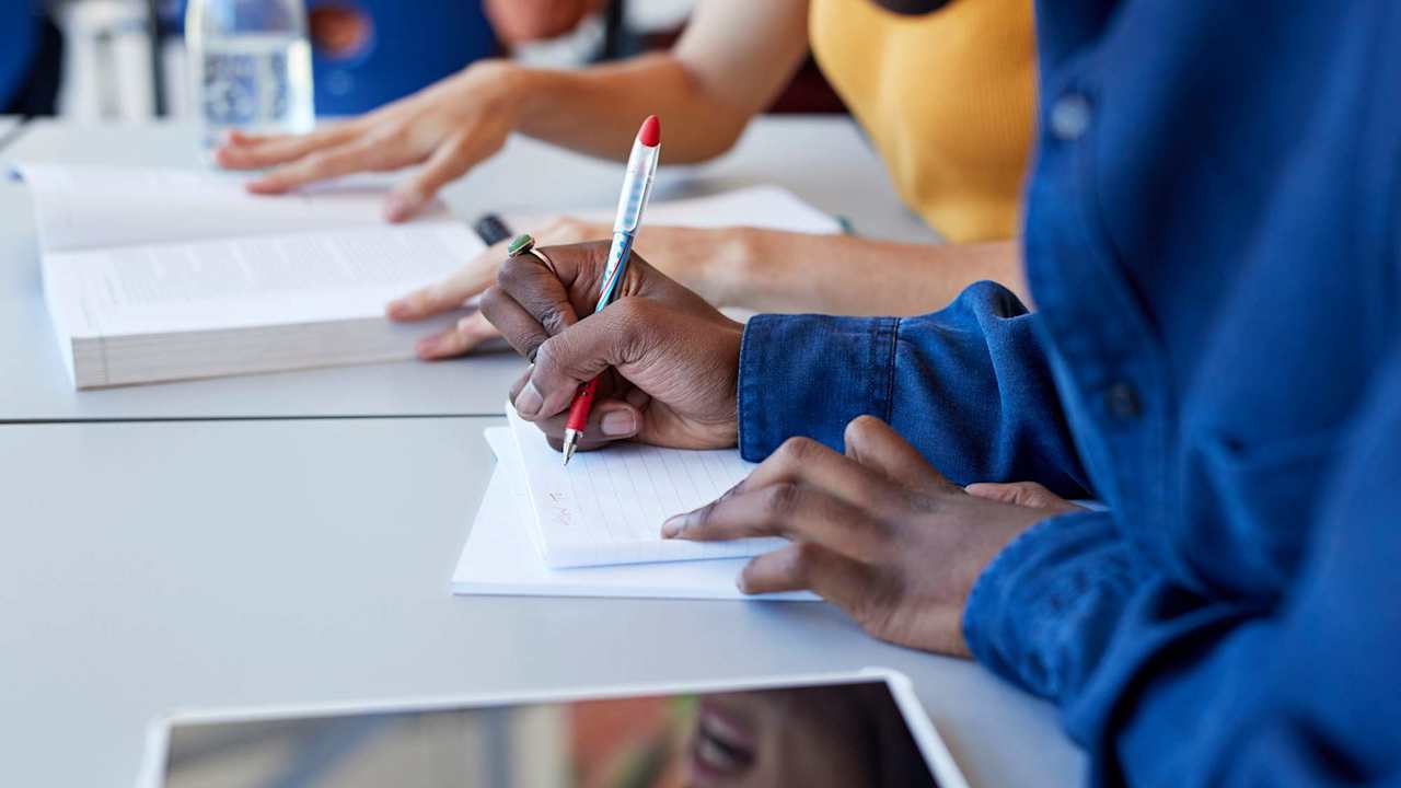 Close up of a Black male high school student writing in a notebook during a study session with a female friend. They are sitting at a table in a high school lunch room and looking at class textbook materials.