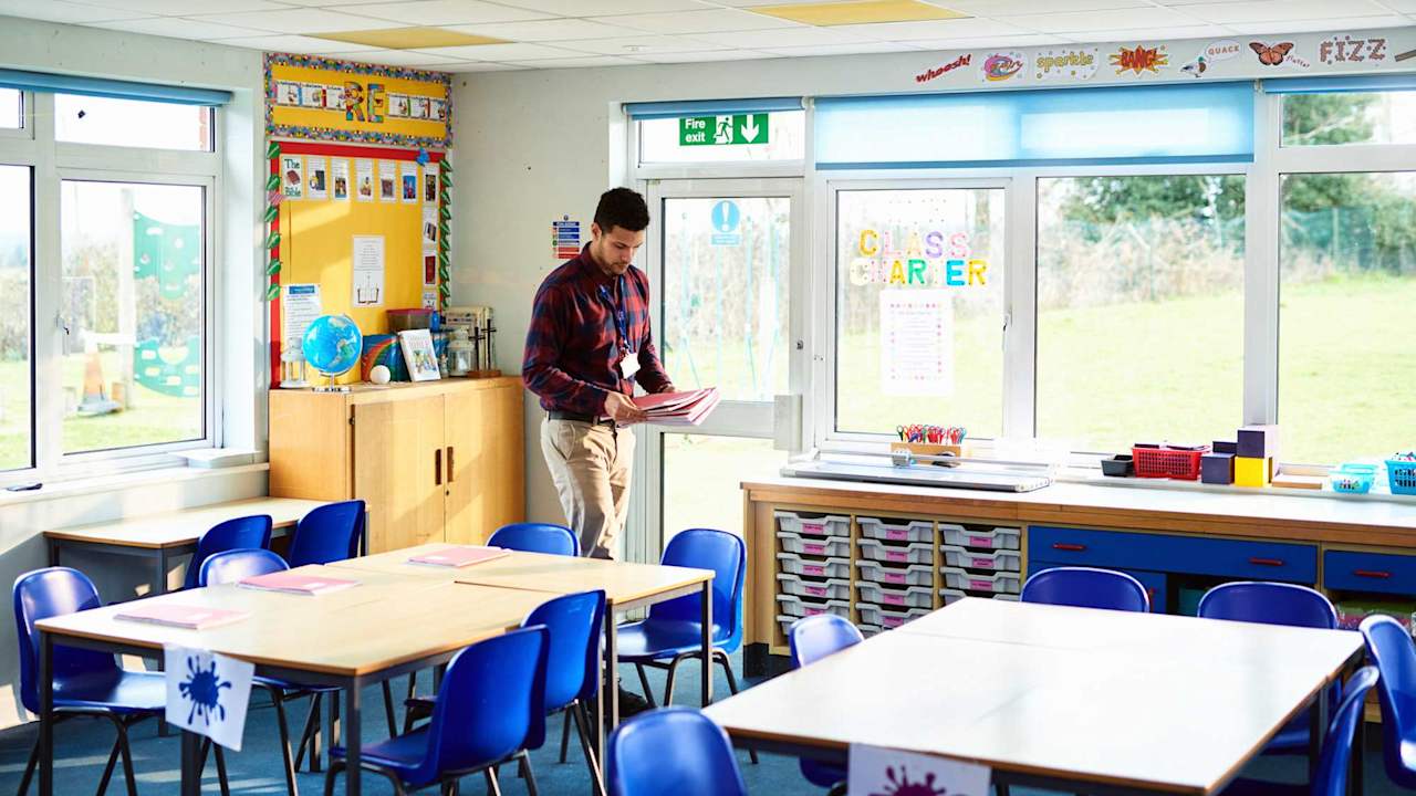 Caucasian male elementary school teacher walking around student desks in a classroom. He is handing out class workbooks before students arrive in the morning.