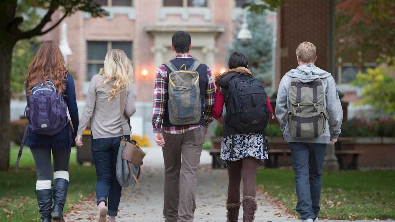 Group of college students walking together on a tree-lined campus path, carrying backpacks and chatting with each other.