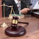 A gavel and the justice scale sit on a desk while a person types on a laptop in the background