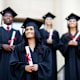 A multicultural group of four college students dressed in graduation gowns and caps stands outside on the steps of a university building with diplomas in their hands.