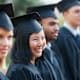 Young college students wearing graduation caps and gowns hold their diplomas while standing in a row and smiling at an outdoor graduation ceremony.