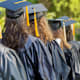 A group of college graduates wearing black caps and gowns stand in line waiting for their diplomas during an outdoor graduation ceremony.