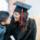 A woman dressed in college graduation regalia leans over and smiles at her young son.