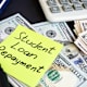 A post-it note with "student loan repayment" written on it laying on top of a stack of U.S. $100 bills.