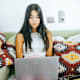 High school senior student types on her laptop while sitting on her living room couch at home.
