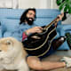 Smiling college student with a prosthetic leg plays a guitar while sitting next to his dog in his apartment living room.
