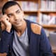 A young man thinks in a library, his head resting in his palm.
