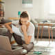 Photo of an anxious college student sitting on the floor in her living room. She has her laptop open and a college tuition bill in her hand.