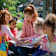 A front view of a teacher and her excited little pupils sitting and doing storytime outdoors in the schoolyard on the benches on a wonderful summer's day.