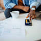 A college student sits in his living room with paper bills and his laptop open on his desk. He is using the calculator app on his mobile phone to go over his finances.