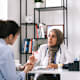 A female Muslim doctor sitting at the desk in her office chats with a female patient during an appointment. The doctor is holding a bottle of prescription medication in her hand and handing it to the patient.