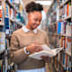 Latina college student standing in an aisle in a university library. She is smiling while reading a textbook for one of her courses.