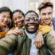 A diverse group of college student friends after class. They are hugging each other and smiling while taking selfie photos.