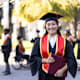 Proud Asian American female college student at her commencement ceremony. She is wearing a black cap and gown and red sash, and is smiling as she holds her degree in her hands.