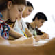 A group of high school students sitting at tables in a classroom. They are writing answers on a paper exam.