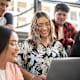 A Latina college student sits with her friends in a university classroom. She is smiling while showing them something on her laptop.