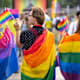 A group of three college students wrapped in bisexual and gay pride flags are waving small flags while watching a gay pride parade during Pride Month.