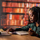 Black female student sitting at a table at the library, using laptop and writing notes while studying for exam.