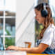 Female Asian American college student sitting at an outdoor table on campus on a sunny warm day. She is wearing headphones and typing on her laptop.