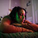 Black female college student lying on her bed at home with the lights off. She is looking off into the distance with a depressed look on her face.