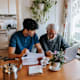 Asian American male social worker discussing medical reports with a Caucasian senior male using a laptop at home.