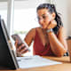 Female student reading on her cell phone. She is sitting at her kitchen table at home, and is referencing materials on her tablet device in front of her.